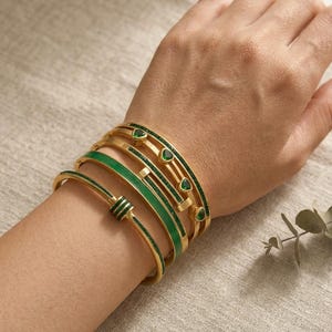 May include: A set of four gold-toned metal bangles worn on a wrist. The bracelets feature green enamel accents, green glitter-textured bands, and small triangular green stones set into the metal. The designs include a smooth band and a hinged clasp.