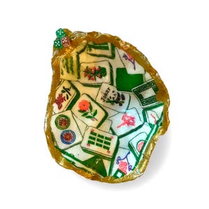 May include: A decorative oyster shell dish featuring a decoupage interior with various mahjong tile designs, including floral patterns and a Joker tile. The rim is painted in a metallic gold finish, and two small dice are attached to the top edge.
