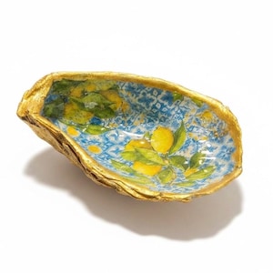 May include: A decorative oyster shell dish featuring a vibrant interior pattern of yellow lemons and green leaves set against a blue and white geometric tile background. The outer edges of the shell are finished with a metallic gold painted trim.