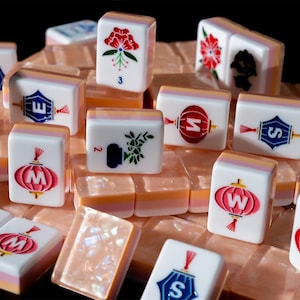 May include: A collection of white Mahjong tiles featuring colorful illustrations of red lanterns with letters, floral designs, and the word joker. The tiles have a translucent, pearlescent base with pink and orange layers, arranged in a scattered pile.