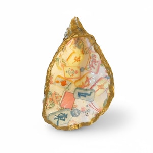 Può includere: Un piattino a forma di conchiglia di ostrica con bordo dipinto in oro e interno in decoupage con un motivo di tessere del mahjong colorate. Le tessere mostrano vari simboli, caratteri e motivi floreali in tonalità di blu, rosso, arancione e verde su sfondo bianco.