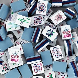 Puede incluir: Una colección de fichas de Mahjong rectangulares con una mezcla de dorsos azules y caras blancas. Las caras blancas muestran varias ilustraciones coloridas, incluyendo tallos de bambú, patrones florales, diseños circulares y máscaras de Joker rojas.