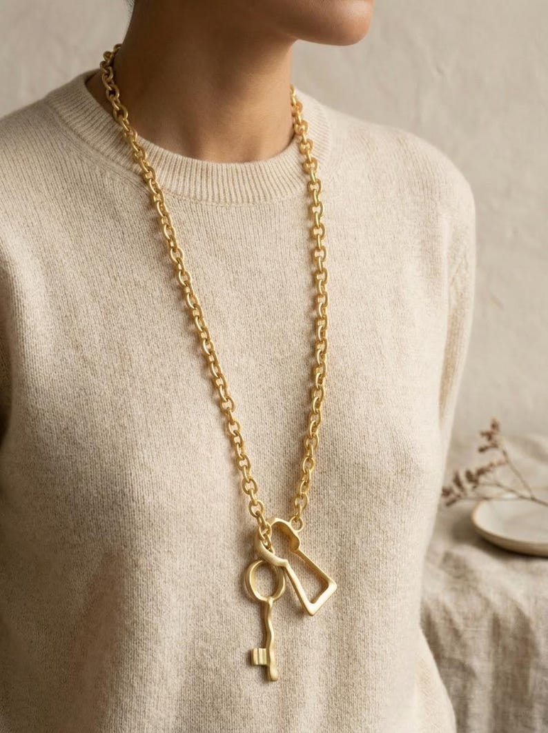 May include: A long gold-toned necklace featuring a thick, chunky chain link design. The pendant consists of two distinct metal charms: a classic skeleton key shape and a stylized keyhole silhouette, both finished in a matte gold-toned metal.