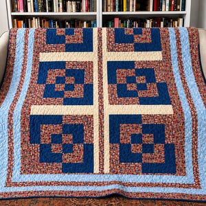 May include: A quilt featuring a geometric pattern of dark blue squares and cream-colored sashing strips. The design incorporates a floral print fabric with small red, blue, and yellow flowers, framed by a light blue border and a floral outer edge.