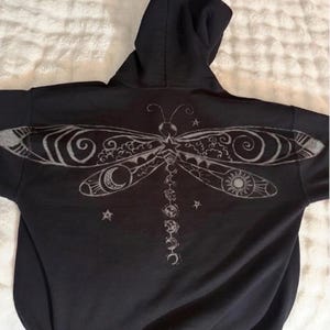 Peut inclure: Un sweat-shirt à capuche noir présentant un grand motif blanc détaillé au dos. Le dessin représente une libellule stylisée avec des motifs complexes dans ses ailes, incluant un croissant de lune, un soleil et une série de phases lunaires.