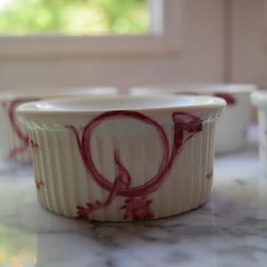 May include: A collection of off-white, ribbed ramekins with a burgundy horn design. The ramekins are arranged on a marble surface, with a blurred background of a window and greenery. These small, round dishes are ideal for baking and serving individual portions.