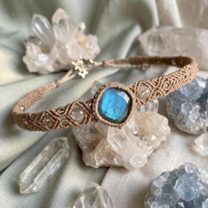 May include: A macrame bracelet featuring a central square faceted labradorite gemstone with a blue iridescent flash, flanked by smaller clear round beads, all set within a tan woven cord band with intricate geometric knot patterns.