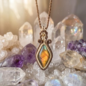 Puede incluir: Un collar con colgante de macramé que presenta una piedra de labradorita en forma de diamante con iridiscencia naranja y amarilla. La piedra está envuelta en un intrincado nudo de cordón marrón y crema, suspendido de un cordón trenzado marrón.