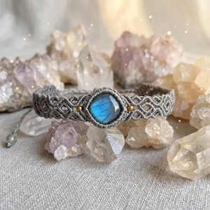 Può includere: Un braccialetto in macramè grigio con una pietra di labradorite centrale a taglio quadrato con un riflesso blu iridescente. Il cinturino in corda intrecciata include piccole perline di metallo dorato su entrambi i lati della pietra e termina con cordini regolabili.