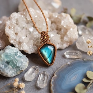 May include: A pendant featuring a faceted, iridescent blue labradorite gemstone encased in brown macrame cord. The setting includes a decorative knot pattern with small brass beads, suspended from a matching brown cord necklace.