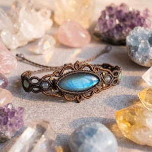 May include: A brown macrame bracelet featuring a central marquise-shaped labradorite gemstone with a blue iridescent flash. The woven cord design includes smaller round beads and intricate knotted patterns with adjustable pull strings at the ends.