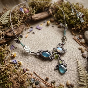 Può includere: Una collana in macramè con una pietra preziosa ovale blu centrale, una pietra di luna iridescente più piccola e una pietra blu a goccia pendente. Il design include ciondoli a foglia color argento, piccole perline verdi e un cordino marrone scuro.