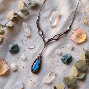 May include: A macrame necklace featuring a teardrop-shaped labradorite pendant with a blue iridescent flash. The brown cord is intricately knotted and accented with small, round, translucent moonstone beads and tiny metallic silver-toned spacers.