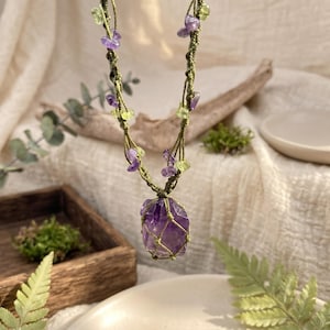 May include: A necklace featuring a large, raw purple amethyst pendant encased in a green macrame cord net. The cord continues upward, decorated with small, irregular purple amethyst and light green peridot stone chips knotted into the string.