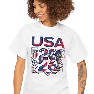 May include: White t-shirt with the word "USA" in navy blue and red block letters. The shirt features a graphic with the numbers "26" and a soccer ball, along with a trophy. The design is in red, white, and blue.