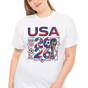 May include: White t-shirt with a graphic design featuring the text "USA 26" in red and blue, along with a soccer ball, a trophy, and the USA flag colors. The shirt is a crew neck style.