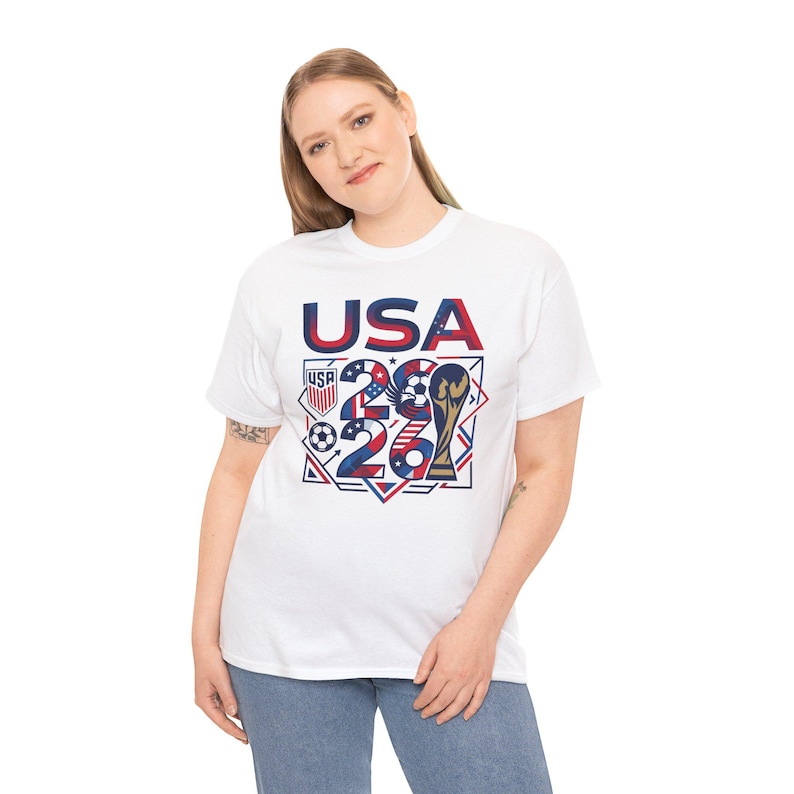 May include: White t-shirt with a graphic design featuring the text "USA 26" in red and blue, along with a soccer ball, a trophy, and the USA flag colors. The shirt is a crew neck style.