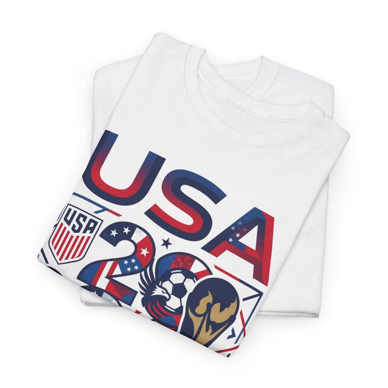 May include: Two white t-shirts are stacked, with the top shirt featuring a graphic design. The design includes the word "USA" in red and blue, along with the number "20" and other patriotic symbols. A soccer ball and a golden trophy are also included.