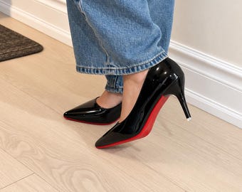 May include: A pair of black patent leather pumps with pointed toes and thin stiletto heels. The soles of the shoes are a vibrant, solid red color. The shoes are worn with blue denim jeans that fall just above the ankle, showing the sleek design.