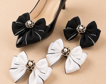 May include: A black patent leather pointed-toe pump featuring a removable black fabric bow with white trim and a central gold-tone flower emblem with a faux pearl. Three additional detachable bows in black and white are displayed alongside the shoe.