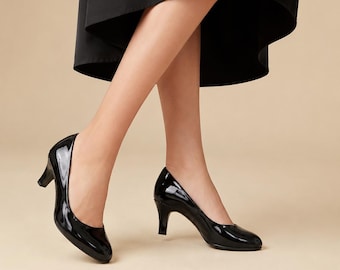 May include: A pair of black patent leather pumps with a rounded toe and a mid-height block heel. The shoes feature a glossy, reflective finish and a simple, classic silhouette. The footwear is paired with a solid black skirt with a high-low hemline.