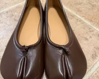 May include: A pair of dark brown leather tabi flats featuring a signature split-toe design, a smooth finish, and a small decorative knot detail at the center of the vamp. The interior is lined with a tan material, and the shoes have a low-profile silhouette.