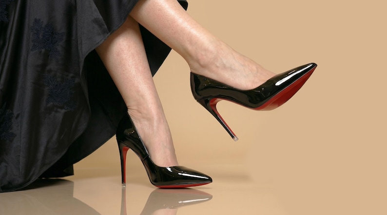 Puede incluir: Un par de zapatos de tac&oacute;n alto de charol negro con punta afilada y tacones de aguja finos. Las suelas de los zapatos son de un color rojo vibrante. Los zapatos se llevan con una falda larga negra con un sutil estampado floral oscuro.