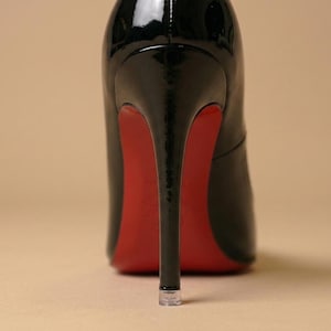 Puede incluir: Un zapato de tac&oacute;n alto de charol negro con acabado brillante, un tac&oacute;n de aguja fino y una suela de color rojo vibrante. El tac&oacute;n tiene una punta de pl&aacute;stico transparente y el zapato se muestra desde una perspectiva trasera directa.