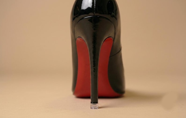 Puede incluir: Un zapato de tac&oacute;n alto de charol negro con acabado brillante, un tac&oacute;n de aguja fino y una suela de color rojo vibrante. El tac&oacute;n tiene una punta de pl&aacute;stico transparente y el zapato se muestra desde una perspectiva trasera directa.