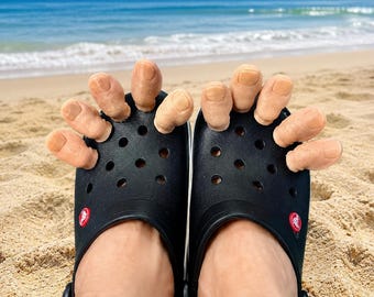 May include: A pair of black clogs featuring a perforated upper design and a small red logo emblem on the side. The footwear is adorned with multiple beige, toe-shaped attachments protruding from the front ventilation holes, creating a unique visual effect.