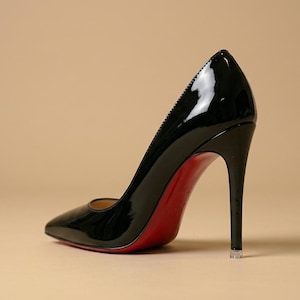Puede incluir: Un zapato de tac&oacute;n de aguja en charol negro con punta afilada y un tac&oacute;n alto y fino. El zapato presenta una suela de color rojo brillante que contrasta con la parte superior negra lustrosa y una peque&ntilde;a tapa protectora transparente en el tac&oacute;n.