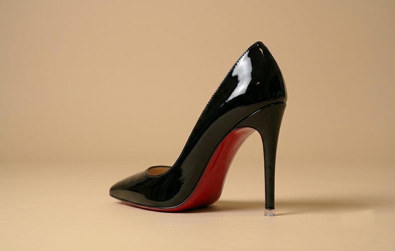 Puede incluir: Un zapato de tac&oacute;n de aguja en charol negro con punta afilada y un tac&oacute;n alto y fino. El zapato presenta una suela de color rojo brillante que contrasta con la parte superior negra lustrosa y una peque&ntilde;a tapa protectora transparente en el tac&oacute;n.