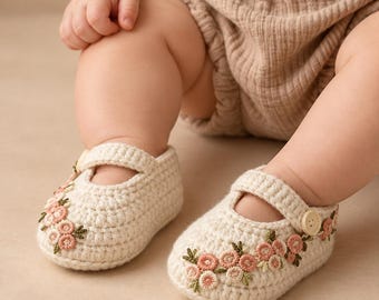 May include: A pair of cream-colored crocheted baby shoes featuring a strap with a small wooden button. The front and sides of the shoes are decorated with embroidered pink flowers and green leaves. The shoes are worn on the feet of a baby.