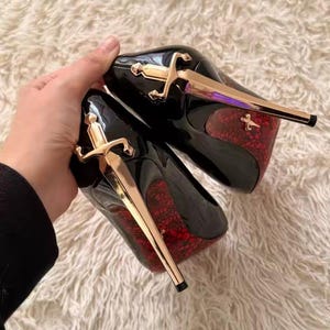 May include: A pair of black high-heeled pumps featuring a gold-toned sword-shaped stiletto heel. The sole of each shoe displays a vibrant red lace-like pattern, and a small gold sword emblem is visible on the red patterned area of the heel.