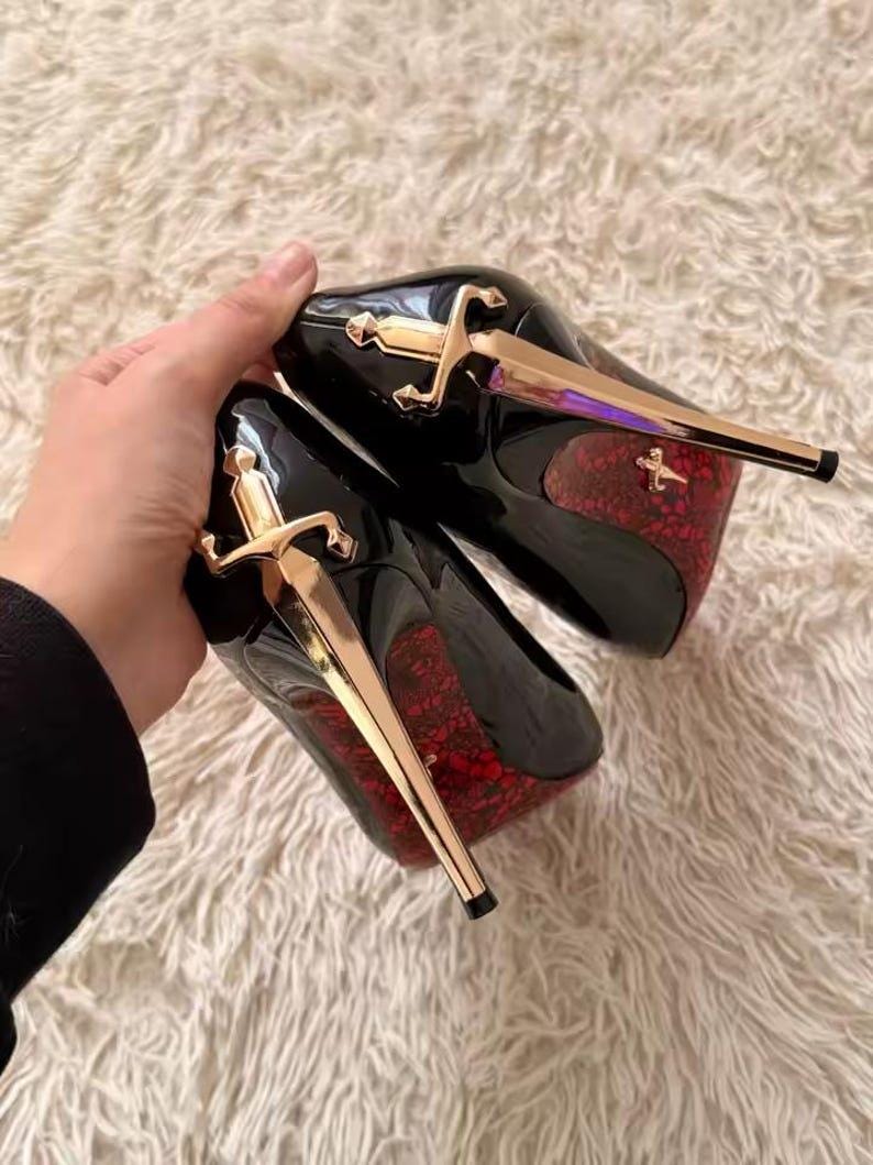 May include: A pair of black high-heeled pumps featuring a gold-toned sword-shaped stiletto heel. The sole of each shoe displays a vibrant red lace-like pattern, and a small gold sword emblem is visible on the red patterned area of the heel.