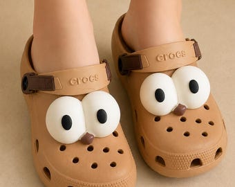 May include: Light brown slip-on clogs featuring a perforated design and a pivoting heel strap with the Crocs logo. Each shoe is decorated with a large, white, cartoon-style eye attachment with a black pupil and a small brown nose detail on the front.