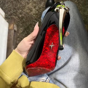 May include: A black patent leather high-heeled shoe featuring a metallic gold stiletto heel and a vibrant red sole with a black lace pattern overlay. A small gold-toned emblem shaped like a dagger is centered on the red sole of the shoe.