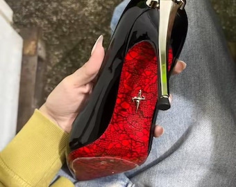 May include: A black patent leather high-heeled shoe featuring a metallic gold stiletto heel and a vibrant red sole with a black lace pattern overlay. A small gold-toned emblem shaped like a dagger is centered on the red sole of the shoe.