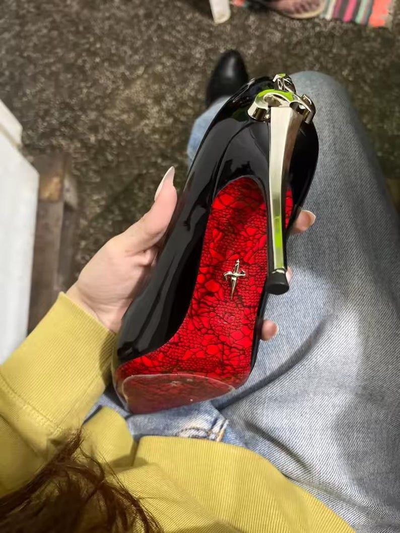 May include: A black patent leather high-heeled shoe featuring a metallic gold stiletto heel and a vibrant red sole with a black lace pattern overlay. A small gold-toned emblem shaped like a dagger is centered on the red sole of the shoe.