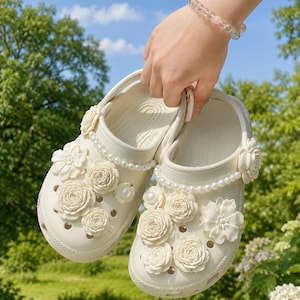 May include: A pair of off-white slip-on clogs featuring decorative embellishments on the upper surface. The shoes are adorned with multiple three-dimensional cream-colored floral charms and a single strand of white faux pearls along the collar.