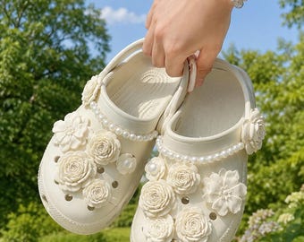 May include: A pair of off-white slip-on clogs featuring decorative embellishments on the upper surface. The shoes are adorned with multiple three-dimensional cream-colored floral charms and a single strand of white faux pearls along the collar.