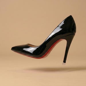 Puede incluir: Un zapato de tac&oacute;n de aguja en charol negro con punta afilada y un tac&oacute;n alto y fino. El zapato presenta un acabado brillante y reflectante en el exterior y una suela roja vibrante visible a lo largo del borde inferior del calzado.