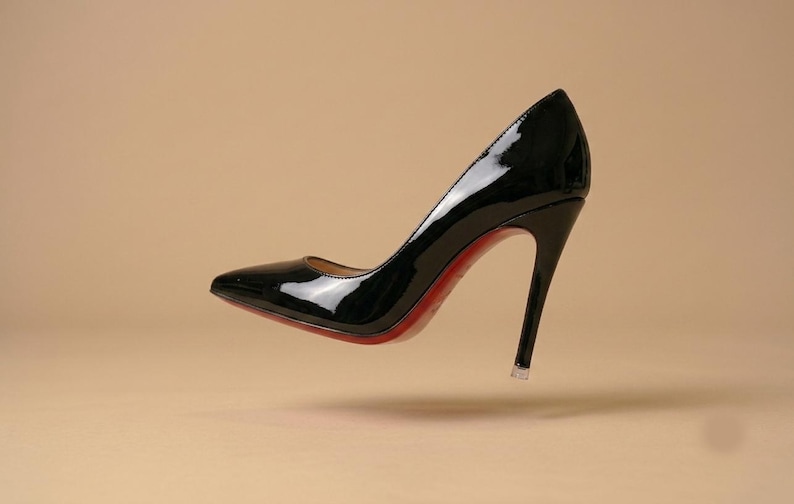 Puede incluir: Un zapato de tac&oacute;n de aguja en charol negro con punta afilada y un tac&oacute;n alto y fino. El zapato presenta un acabado brillante y reflectante en el exterior y una suela roja vibrante visible a lo largo del borde inferior del calzado.