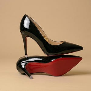 Puede incluir: Un par de zapatos de tac&oacute;n alto de charol negro con punta afilada y tacones de aguja finos. Un zapato est&aacute; de pie mientras que el otro descansa de lado, revelando una suela de color rojo vibrante. El forro interior es de color beige claro.