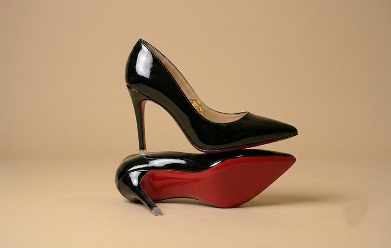 Puede incluir: Un par de zapatos de tac&oacute;n alto de charol negro con punta afilada y tacones de aguja finos. Un zapato est&aacute; de pie mientras que el otro descansa de lado, revelando una suela de color rojo vibrante. El forro interior es de color beige claro.