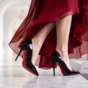 May include: A pair of black patent leather high-heeled pumps with pointed toes and bright red soles. The shoes are worn with a flowing, layered dark red chiffon skirt. A small white card with a gold crown icon and the text Sole Trend is visible.
