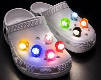 May include: A pair of white slip-on clogs featuring multiple small, round, battery-operated LED lights attached to the top surface. The lights emit various colors including yellow, blue, pink, green, orange, and white, illuminating the shoe surface.