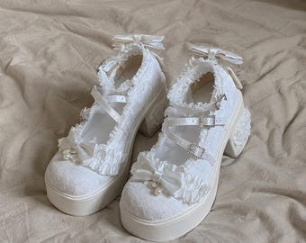 May include: A pair of white platform shoes featuring lace fabric, ruffled trim, and satin bows on the toe and heel. The shoes include multiple straps with silver-toned buckles, decorative pearl accents, and a thick block heel design.