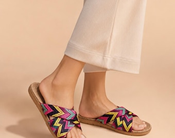 May include: A pair of slide sandals featuring crisscross straps with a woven chevron pattern in vibrant shades of magenta, teal, yellow, and black. The sandals have a flat, textured brown sole and are worn with light-colored ribbed trousers.
