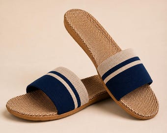 May include: A pair of slide sandals featuring a textured, tan woven footbed and a wide fabric strap. The strap is navy blue with two horizontal beige stripes across the center. The sandals have a flat sole and an open-toe design.